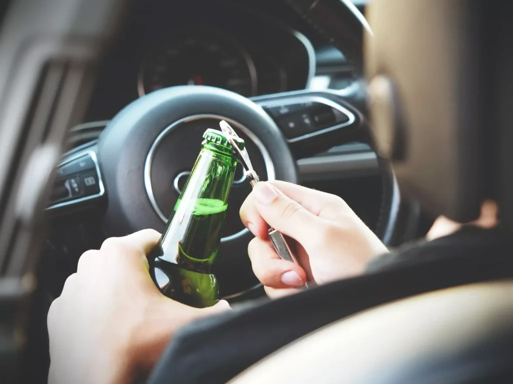 A man opening up a beer bottle while behind the wheel of a car.