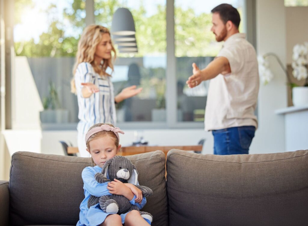 An upset child on the couch while a couple is fighting.