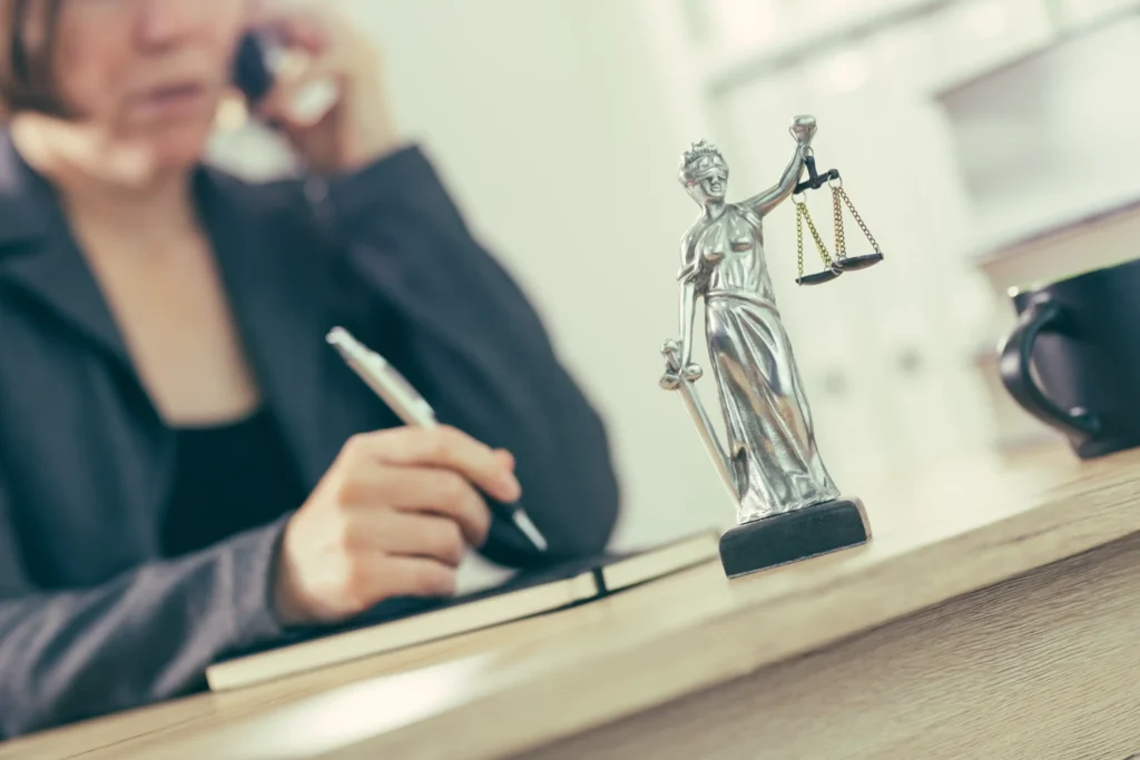 An attorney speaking on the phone while sitting at her desk, holding a pen and justice scales.