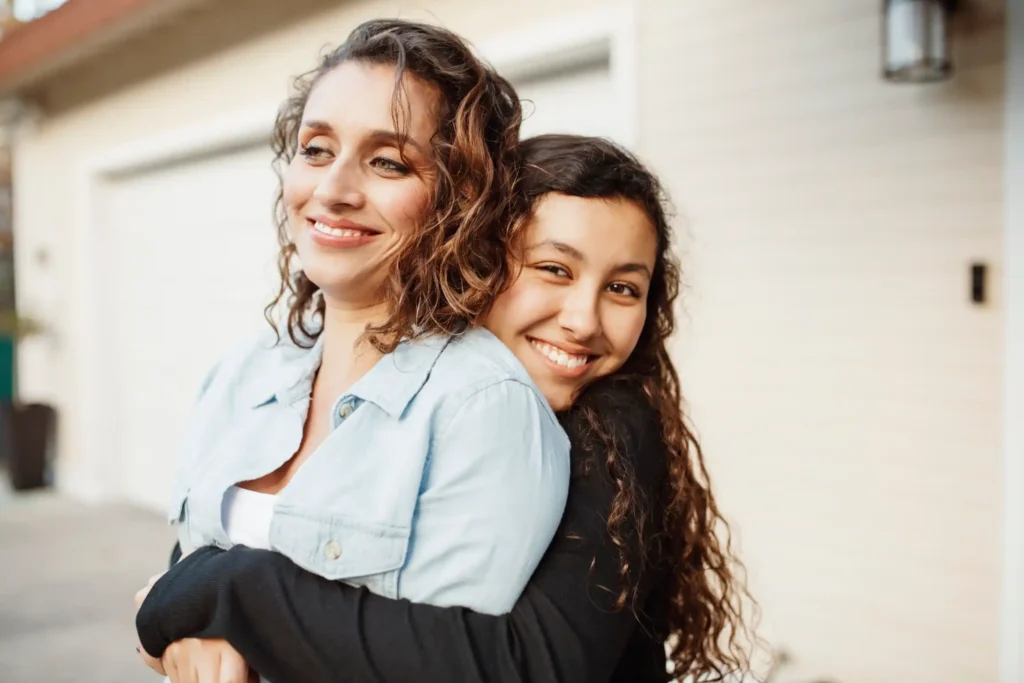 A smiling daughter hugging her smiling mother from behind.