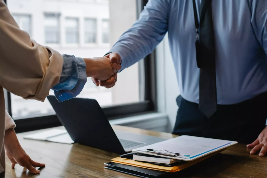 An attorney shaking his client's hand while he is at his desk with a laptop and paperwork.