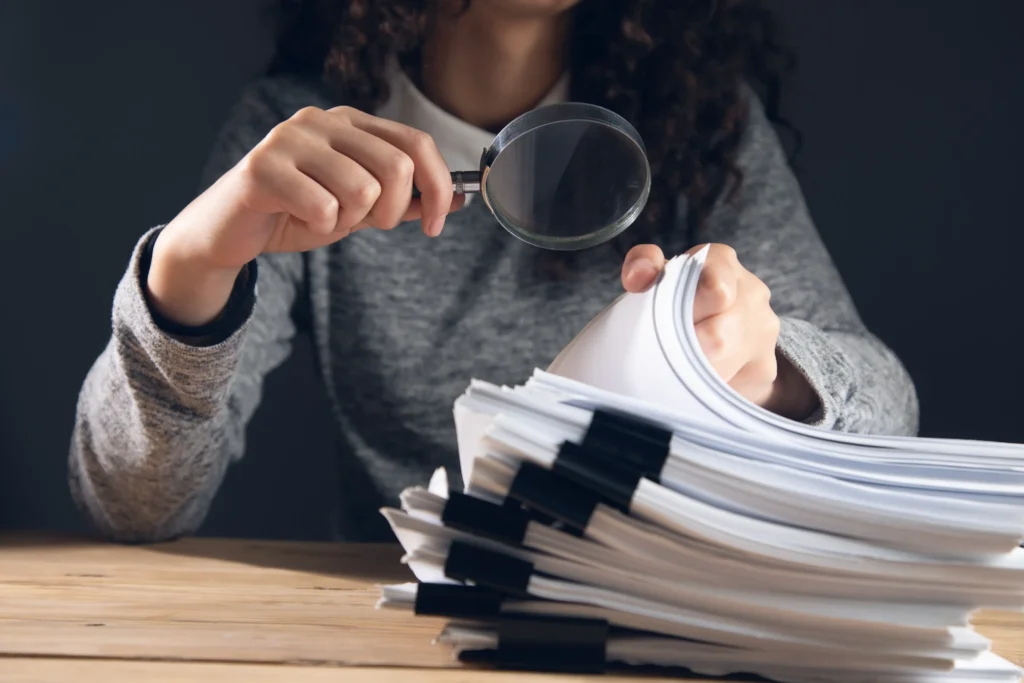 Woman examining a large stack of legal documents with a magnifying glass, symbolizing the detailed investigation and case review involved in defending domestic violence charges in Dallas.